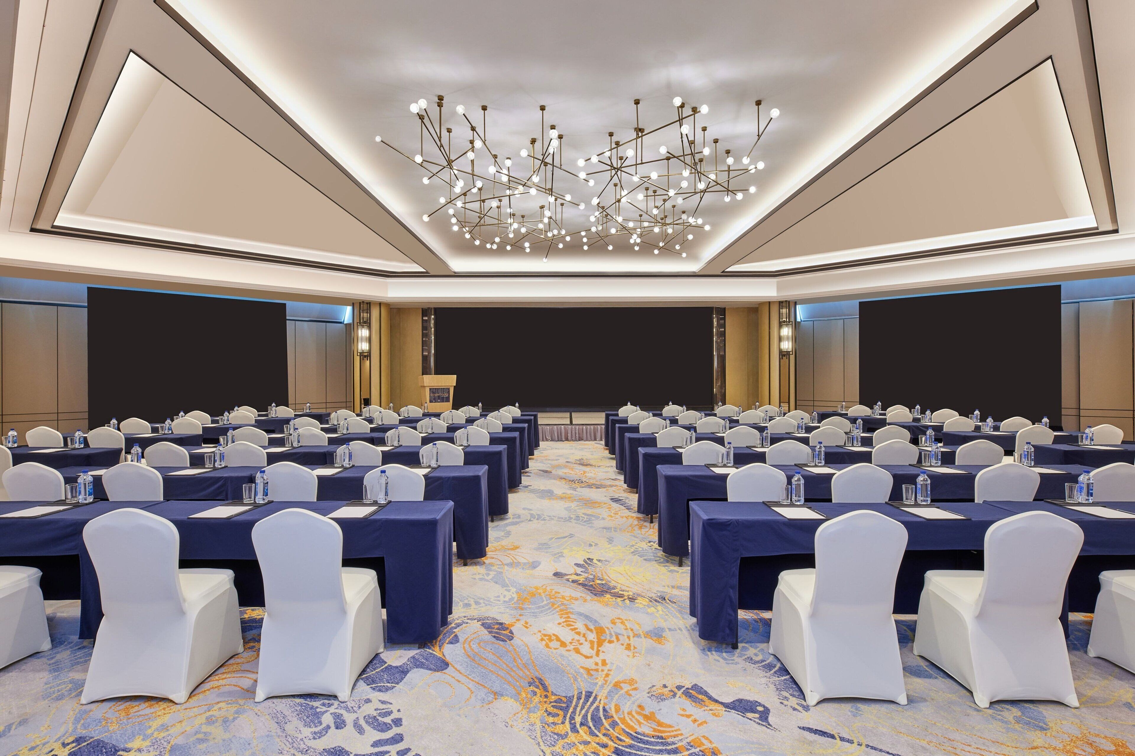 A conference room with rows of tables and chairs, a large chandelier, and a carpeted floor.