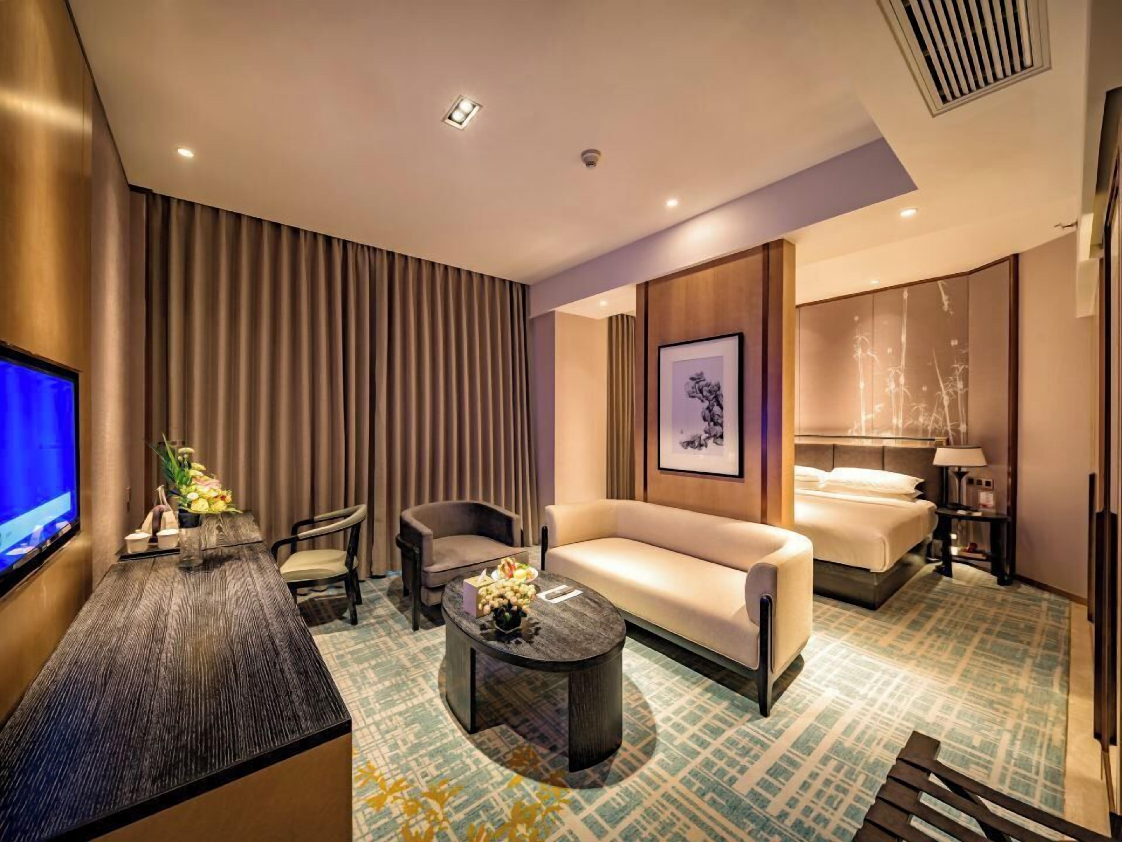 A hotel room with a sofa, two chairs, a coffee table, a TV, a bed, and a painting on the wall.