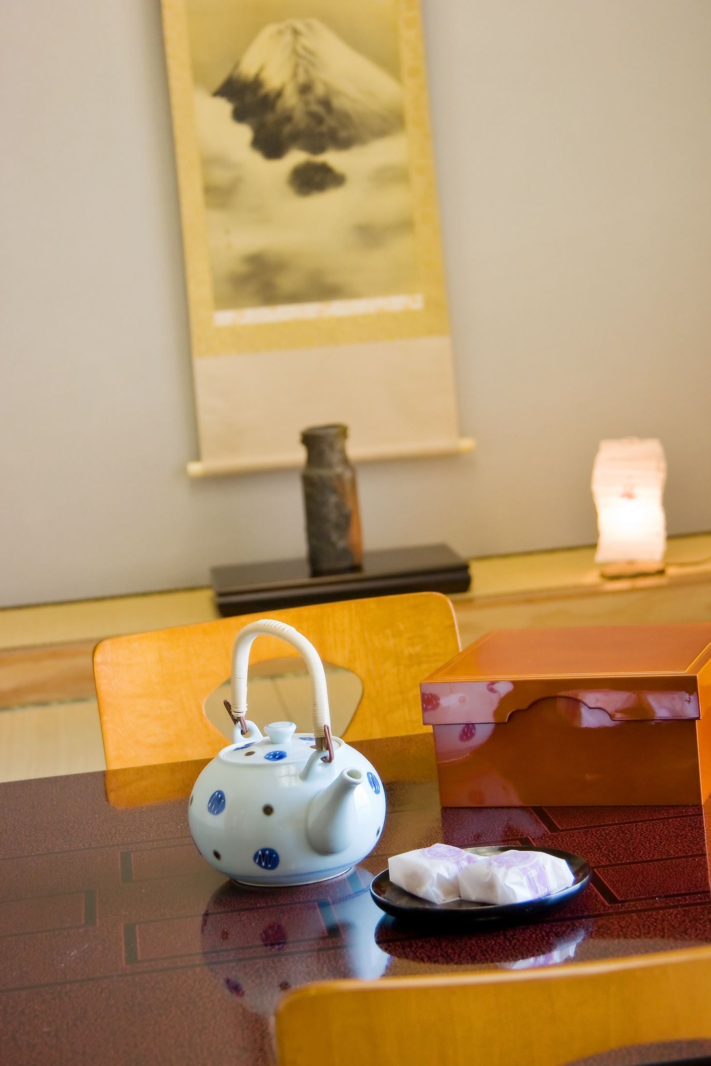 A table with a white teapot with blue patterns, a plate with food, and a framed picture of a mountain on the wall.