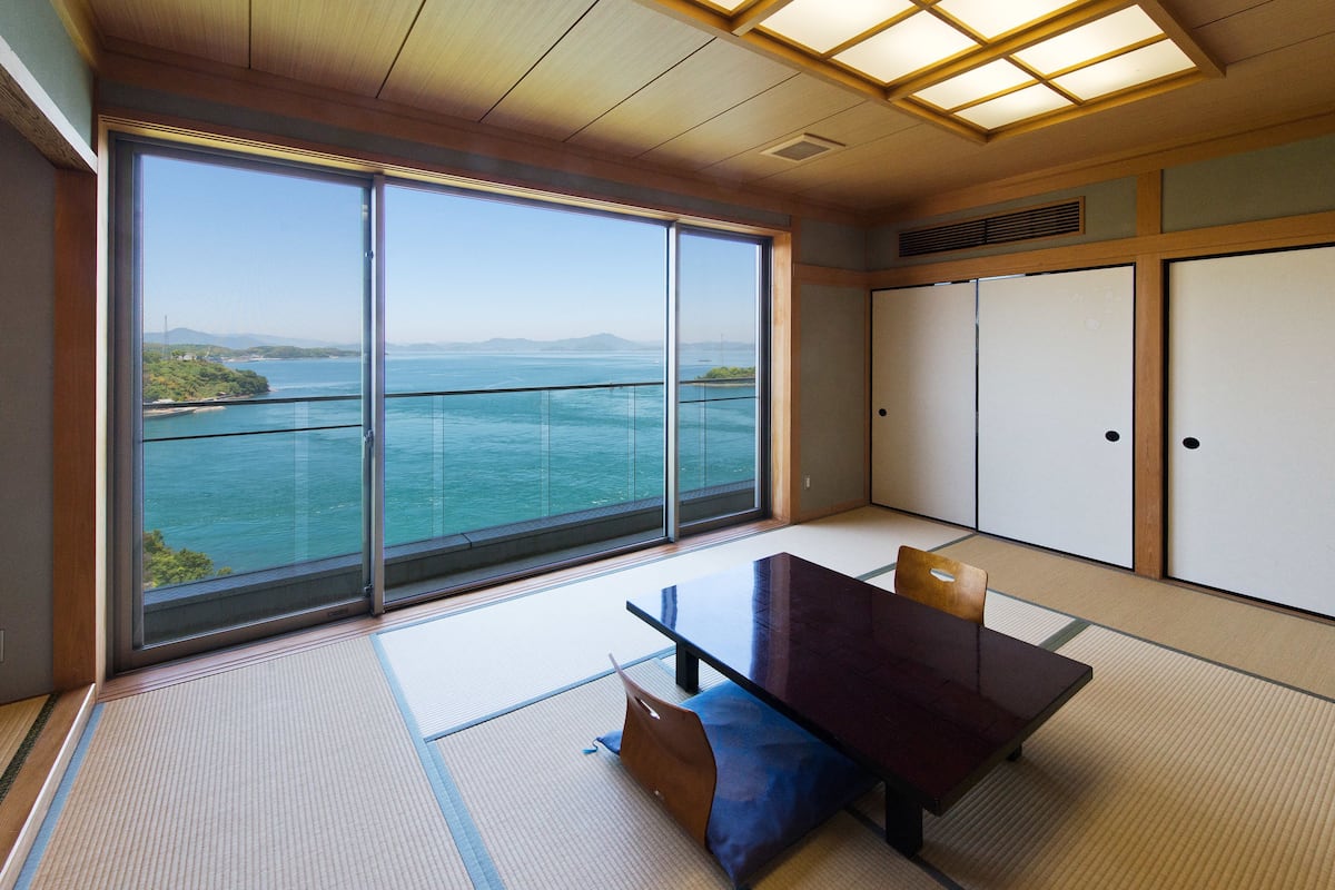 Quarto com piso de tatame, uma mesinha baixa e portas deslizantes. Uma janela ampla oferece vista para o mar e terras distantes.