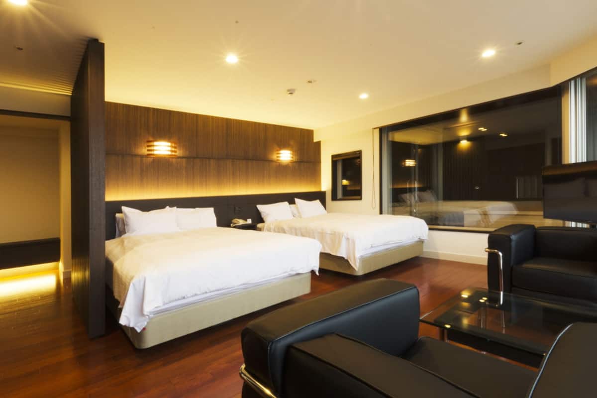A modern hotel room with two beds, a sofa, a glass table, and a large window.