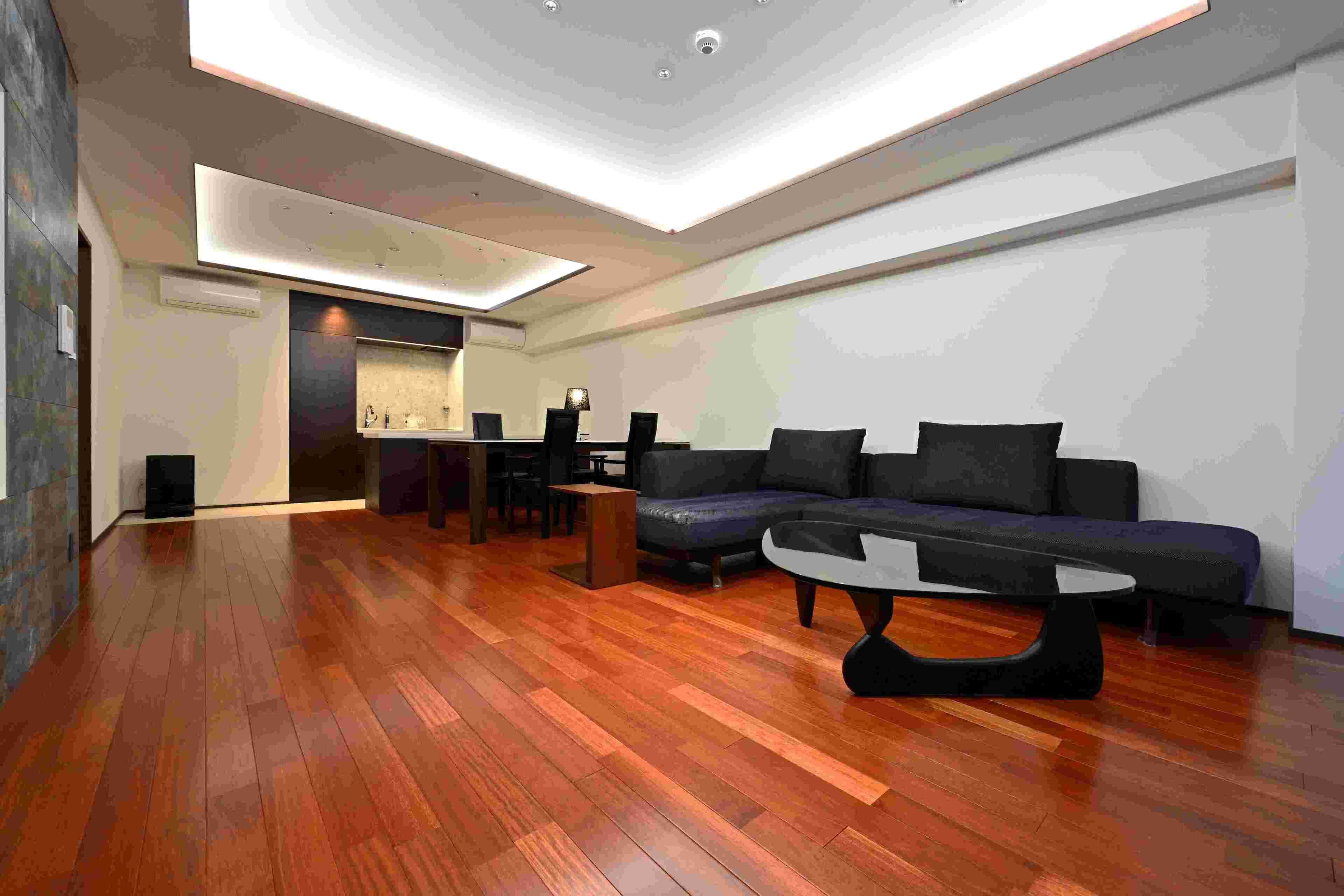 Uma sala de estar moderna com piso de madeira, um sofá preto, uma mesa de centro e uma cozinha ao fundo.