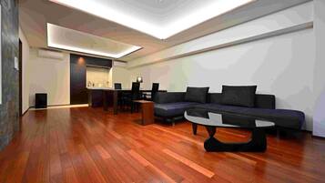 Uma sala de estar moderna com piso de madeira, um sofá preto, uma mesa de centro e uma cozinha ao fundo.