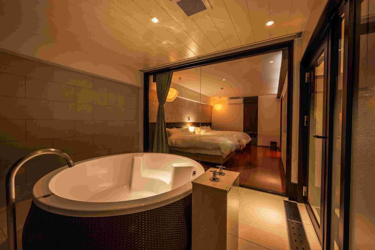 A modern bathroom with a large bathtub, a glass shower enclosure, and a visible bedroom with a bed and nightstand.