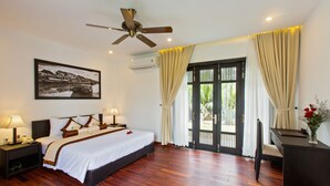 Habitación de hotel con cama, mesitas de noche, escritorio, silla, ventilador de techo y puerta que da a un espacio exterior.