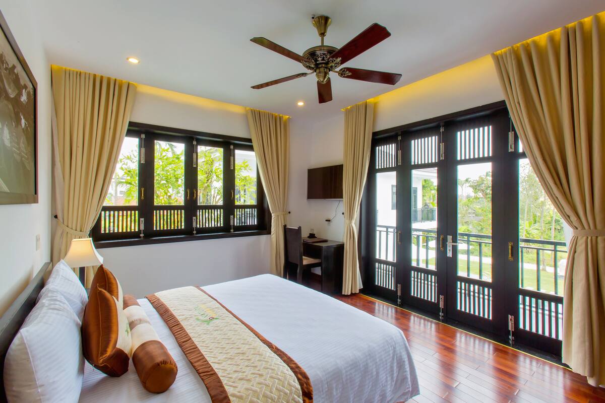A hotel room with a bed, curtains, a ceiling fan, a desk with a chair, and large windows with a view of greenery.