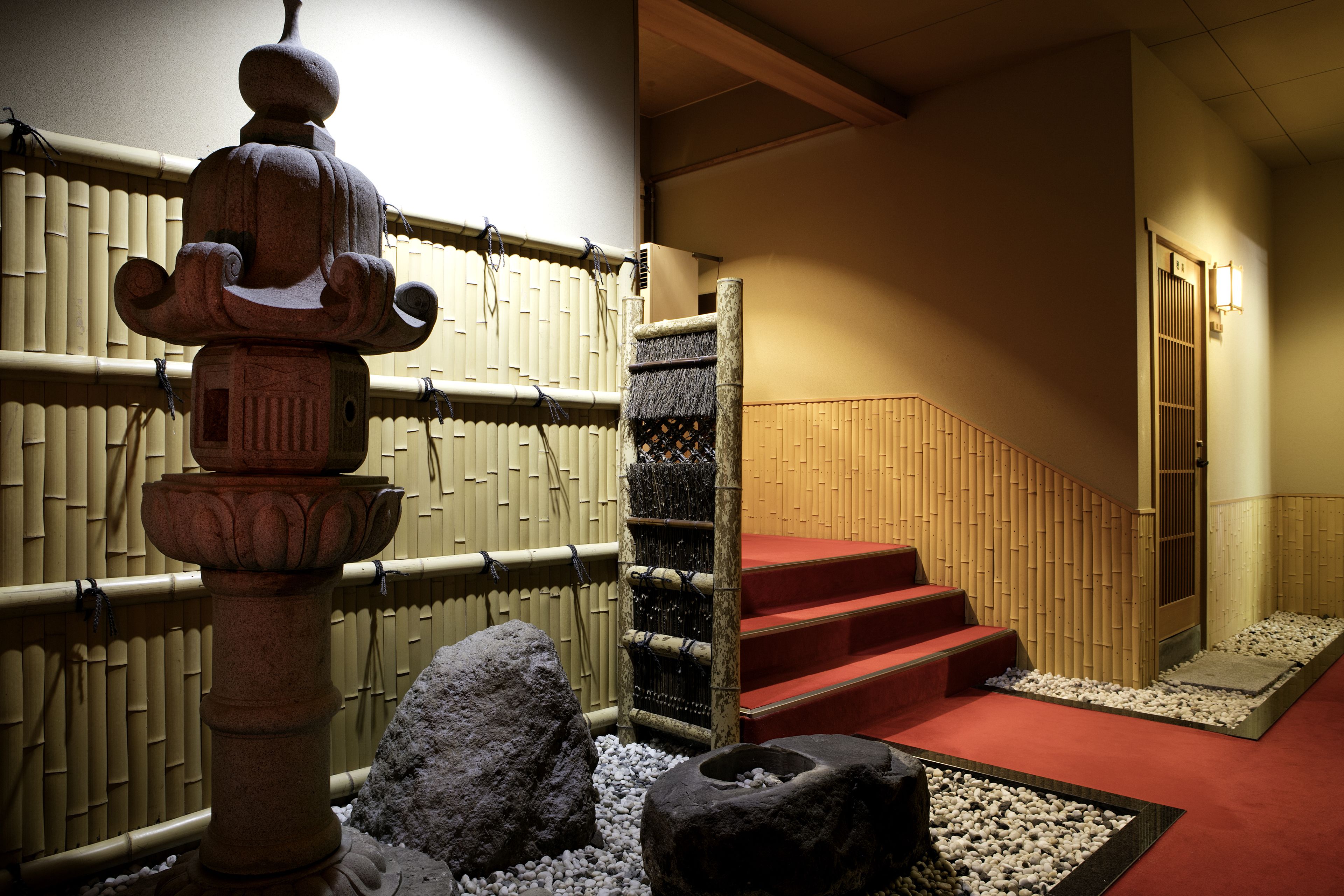 A traditional Japanese interior with a stone water feature, bamboo wall, and a red carpeted staircase.