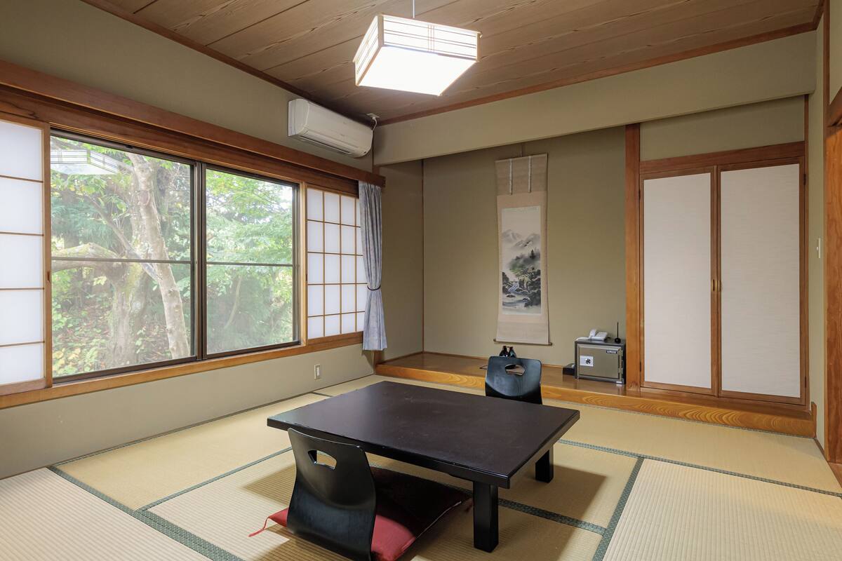 Una habitación japonesa tradicional con suelo de tatami, puertas correderas, una mesa y vistas a árboles.