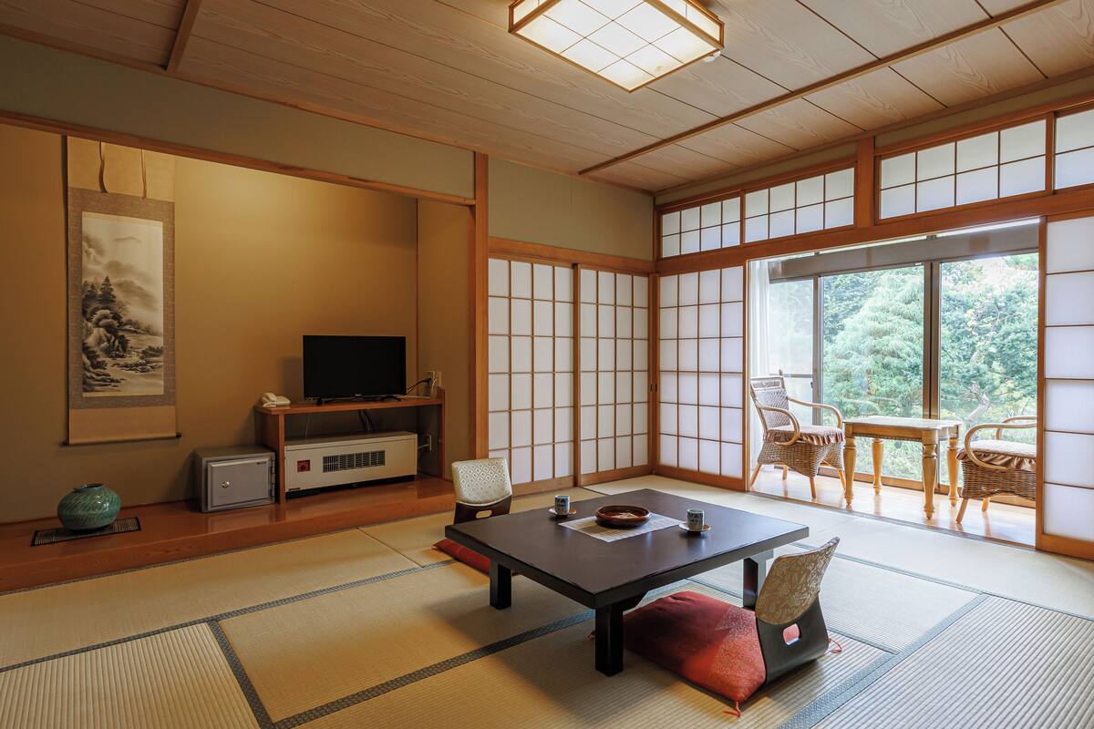 Una habitación japonesa tradicional con suelo de tatami, una mesa baja, un televisor y vistas a zonas verdes a través de grandes ventanales.