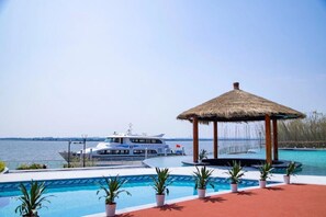 A pool area with a thatched gazebo, a white boat docked nearby, and a view of the water.