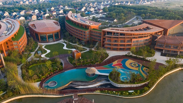 Vue aérienne d’un complexe hôtelier moderne composé de plusieurs bâtiments aux formes courbes, d’une piscine centrale et de jardins aménagés.