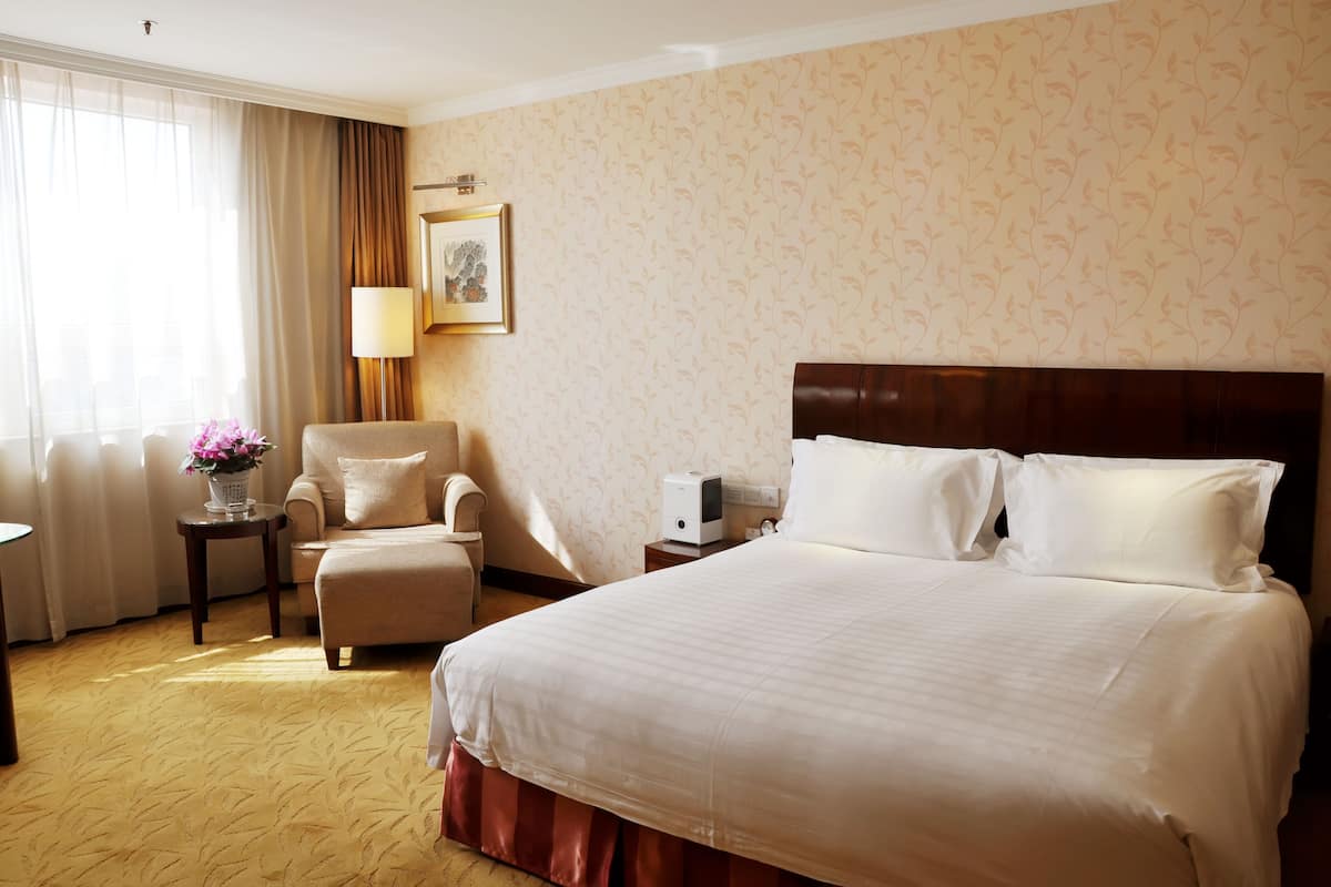 Een hotelkamer met een groot bed, een stoel, een klein tafeltje met een lamp en een raam met gordijnen.