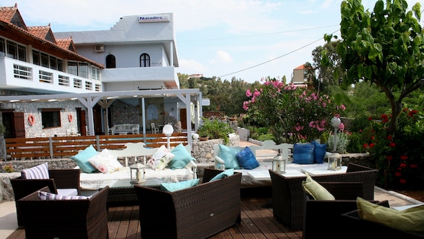 An outdoor patio area with wicker furniture, a stone wall, and a view of a building with a sign reading "Naiades". - Naiades Almiros River Hotel (Agios Nikolaos)