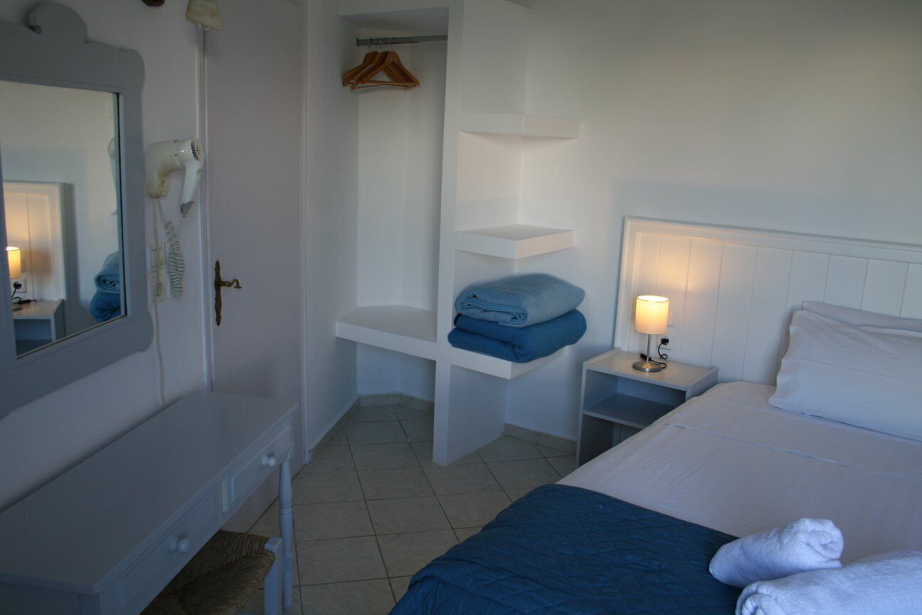 A hotel room with a bed, a vanity with a mirror, a hairdryer, a shelf with folded towels, and a bedside table with a lamp.
