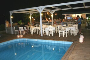 An outdoor pool area with white tables and chairs, surrounded by a wooden deck and a covered seating area with string lights. - Naiades Almiros River Hotel (Agios Nikolaos)
