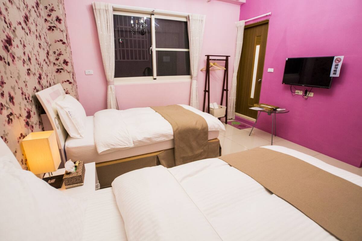 A hotel room with two beds, a television, a window with curtains, a bedside table, and a lamp.