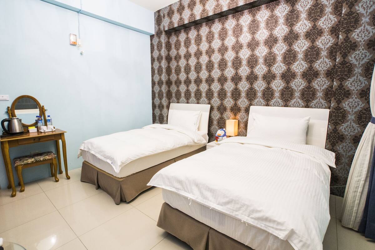 A hotel room with two beds, a small table with a kettle, and a mirror.