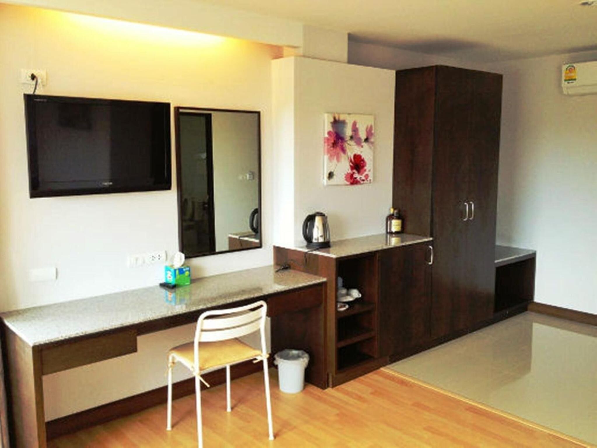 Una habitación pequeña y moderna con escritorio, silla y armario. Hay un televisor de pantalla plana montado en la pared, un estante con una tetera y una botella, y un cesto de basura.