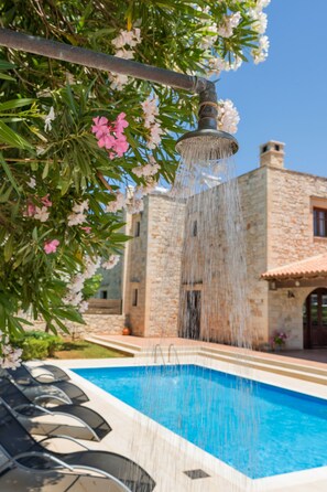 A pool with water cascading from a fountain, surrounded by stone buildings and flowering plants. - Adam villas (Kissamos)