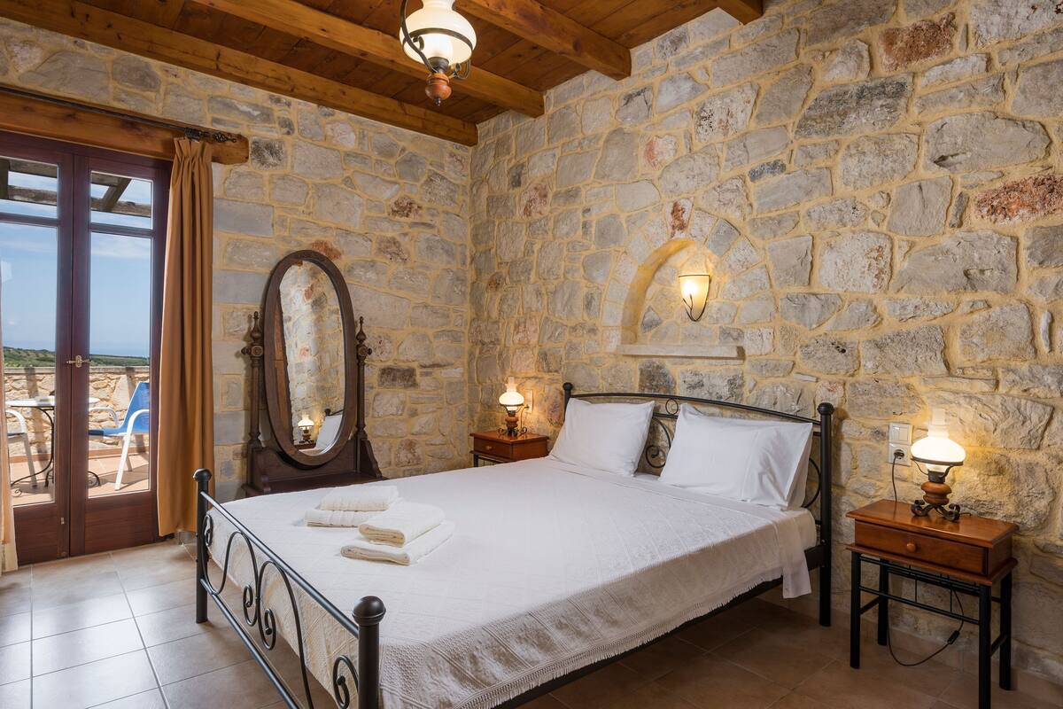 Ein Schlafzimmer mit Steinwänden, einem Bett mit weißer Bettwäsche, einer Holzbalkendecke und Blick durch eine Glastür.
