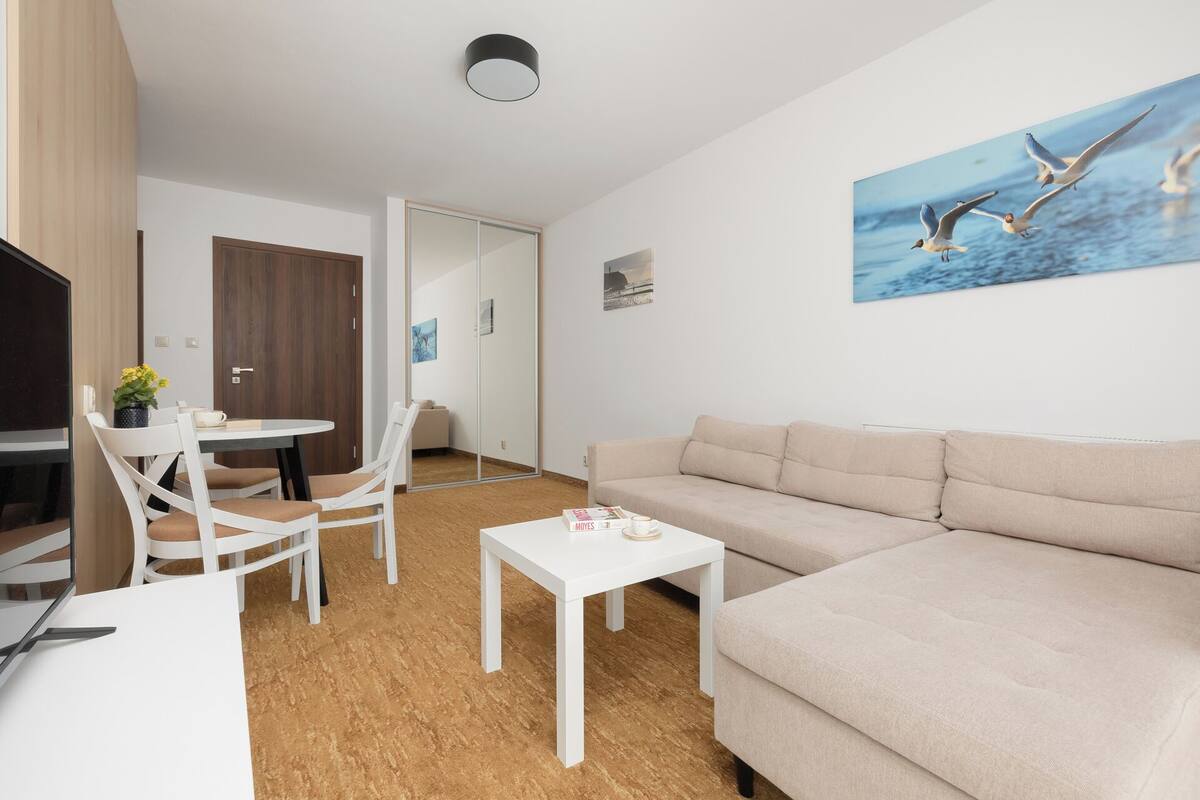 A modern living room with a beige sofa, white chairs, a small white table, and a painting of birds on the wall.