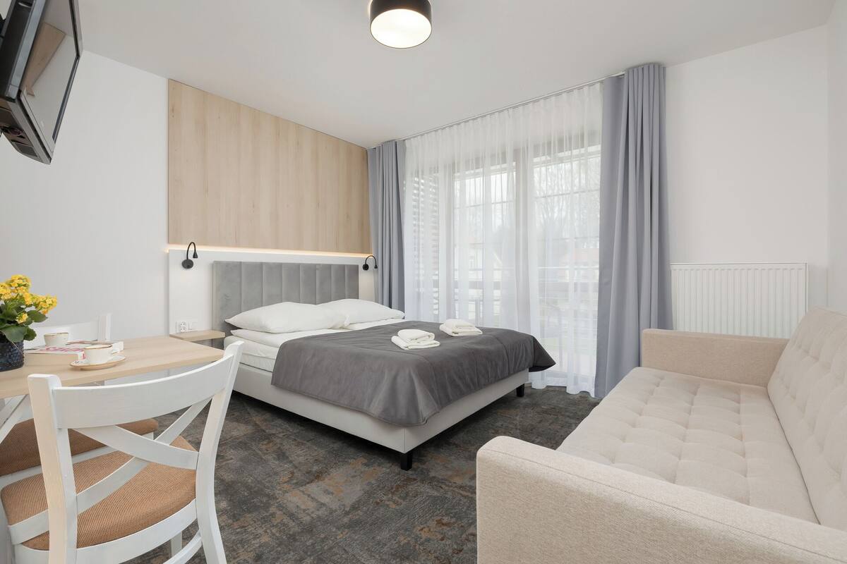 A hotel room with a bed, a sofa, a desk with a chair, a TV, and a window with curtains.