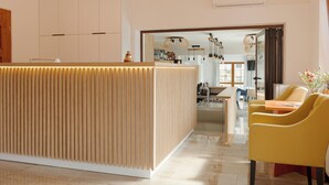 A modern reception area with a wooden reception desk, a dining area with yellow chairs, and a kitchen visible through an open doorway. - Willa Florek (Rewal)