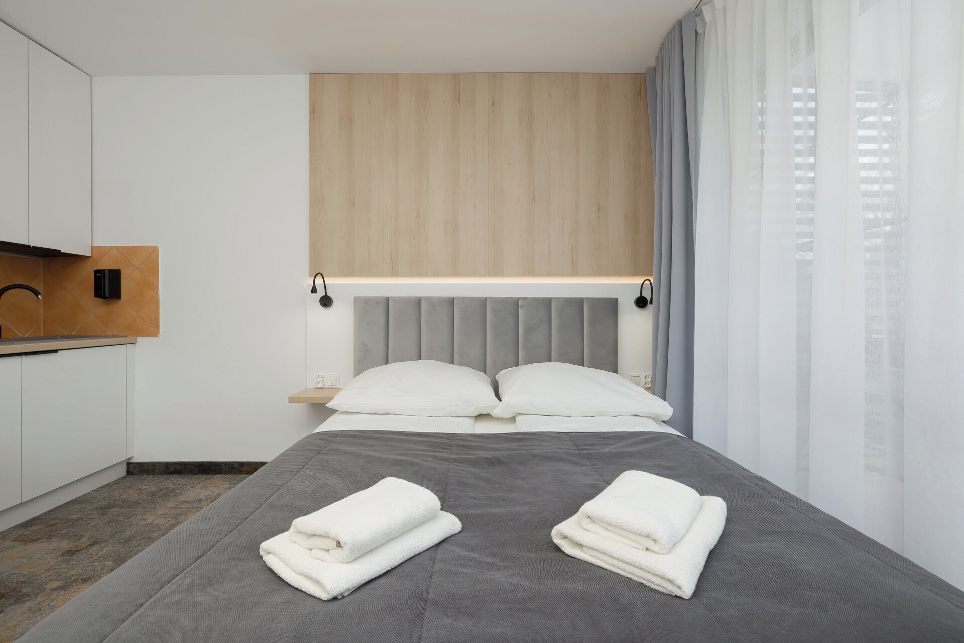 Ein modernes Hotelzimmer mit einem grauen Bett, weißen Kissen und einem hölzernen Kopfteil. Ein kleiner Nachttisch mit einer Lampe und einer Steckdose steht daneben.