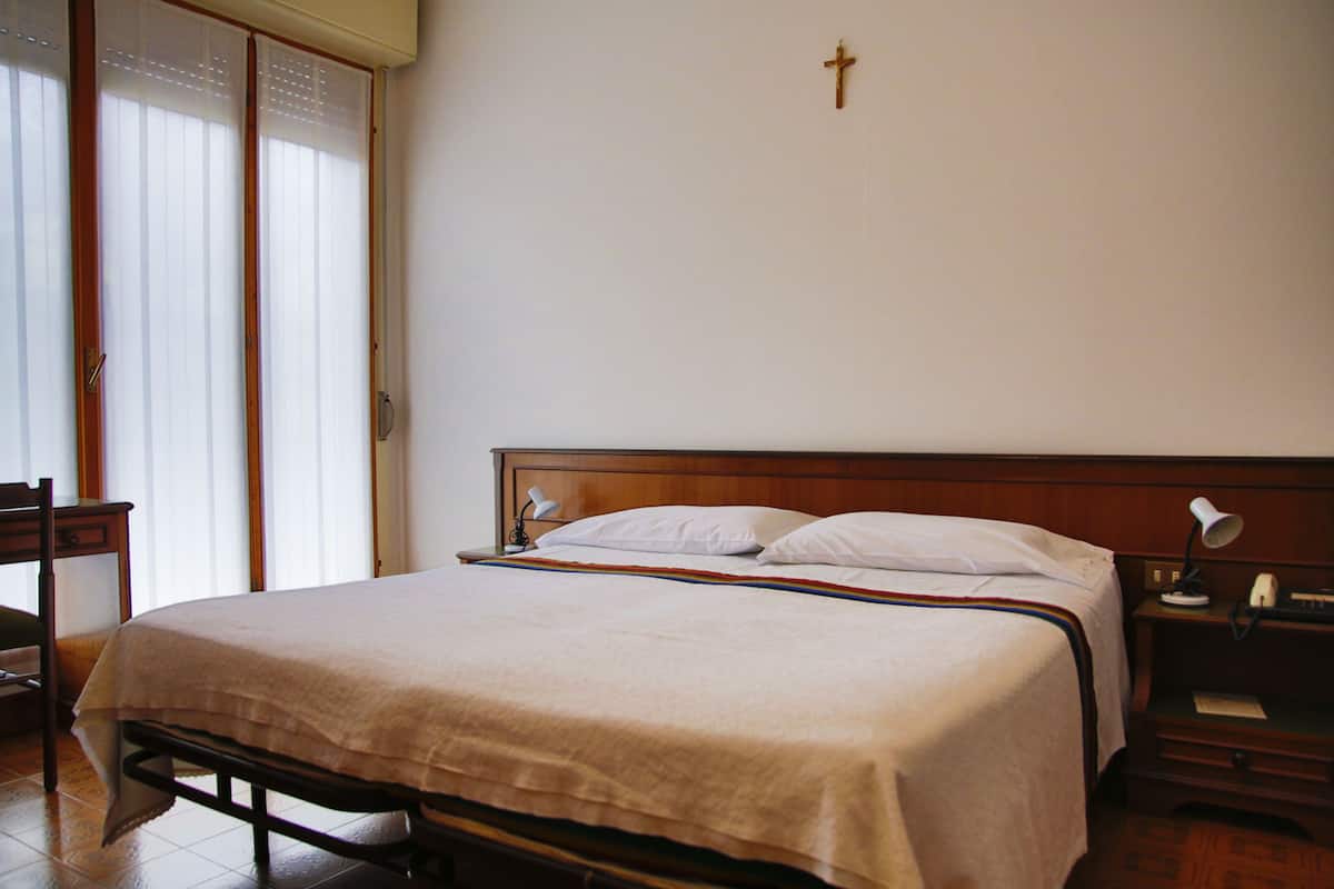 Een slaapkamer met een bed, een nachtkastje, een stoel en een kruis aan de muur.