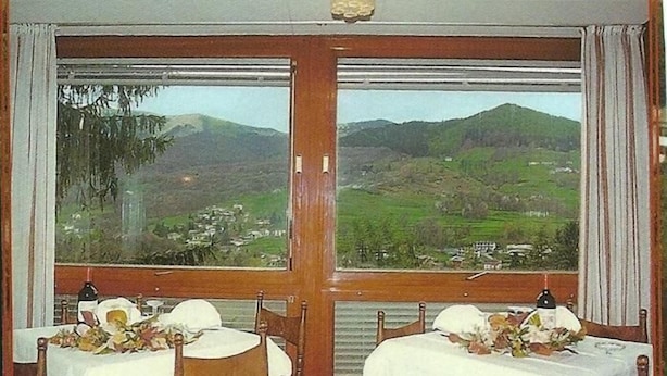 Een eetruimte met twee tafels voor twee personen, elk met een wit tafelkleed en bloemdecoraties. Grote ramen bieden uitzicht op een bergachtig landschap.