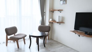 Una sala moderna con dos sillones tapizados grises, una mesita redonda, un televisor montado y estanterías de madera con un jarrón blanco y una planta pequeña.