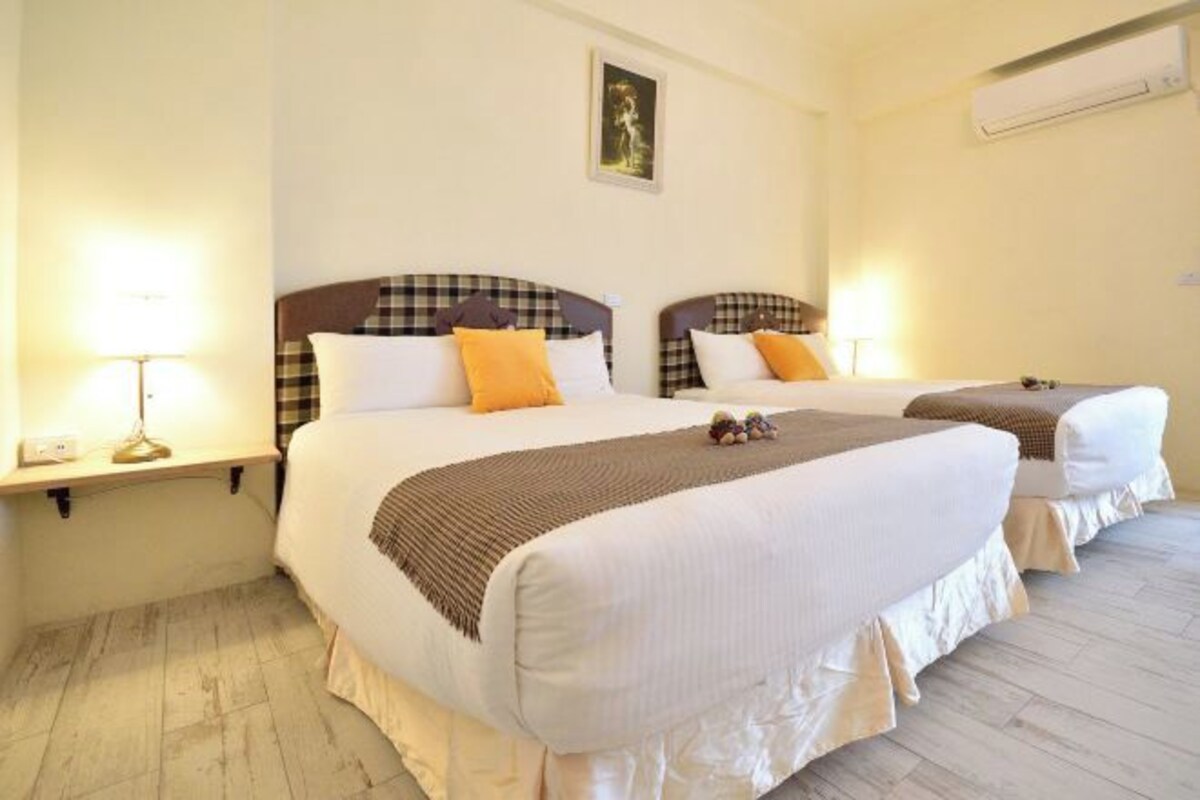 Une chambre d’hôtel avec deux lits, chacun doté d’une tête de lit à carreaux, de draps blancs et d’un plaid marron. Il y a une table de chevet avec une lampe de chaque côté des lits, et un tableau encadré au mur, au-dessus des lits.