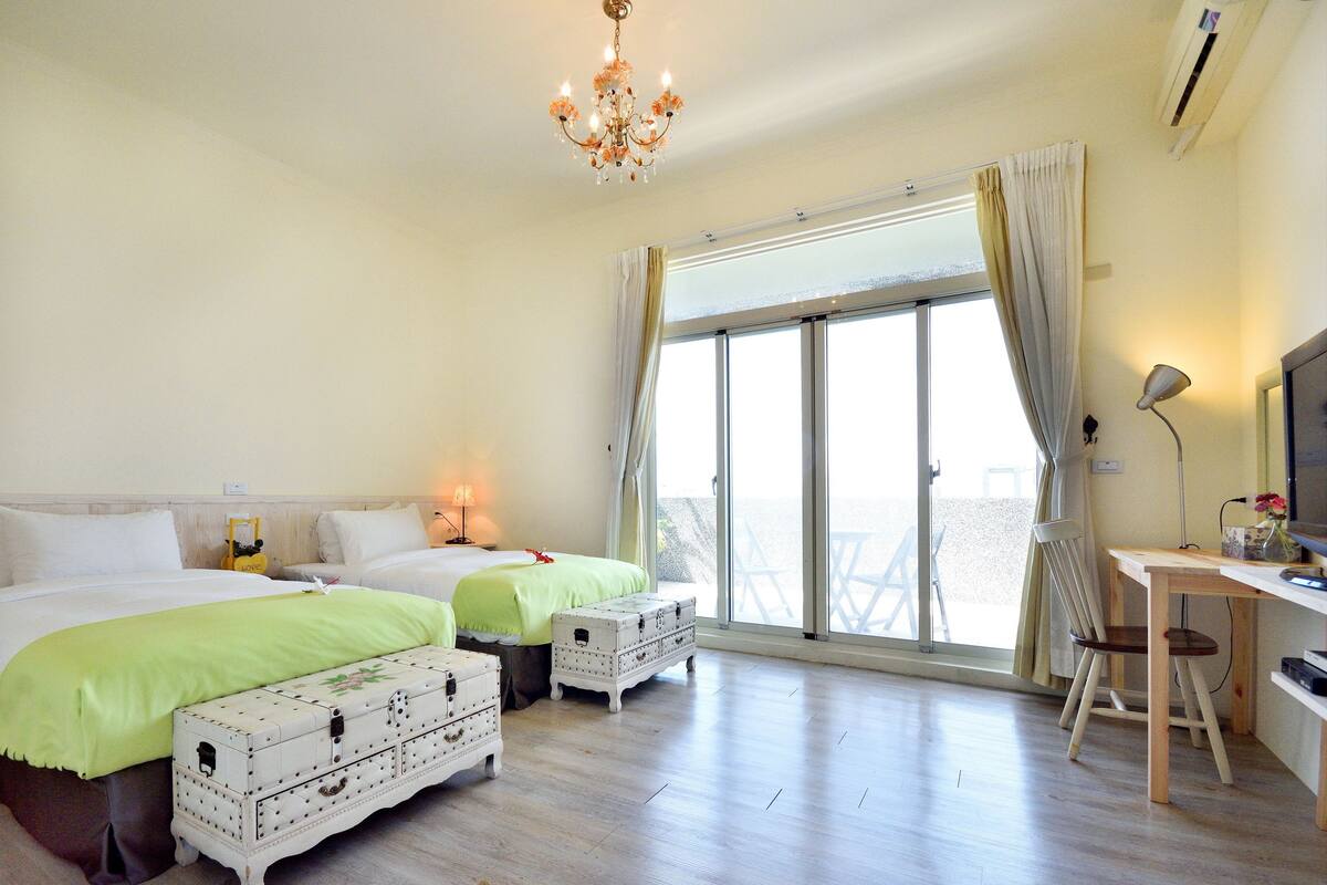 A hotel room with two beds, a desk, a chair, a chandelier, and a large window with curtains.