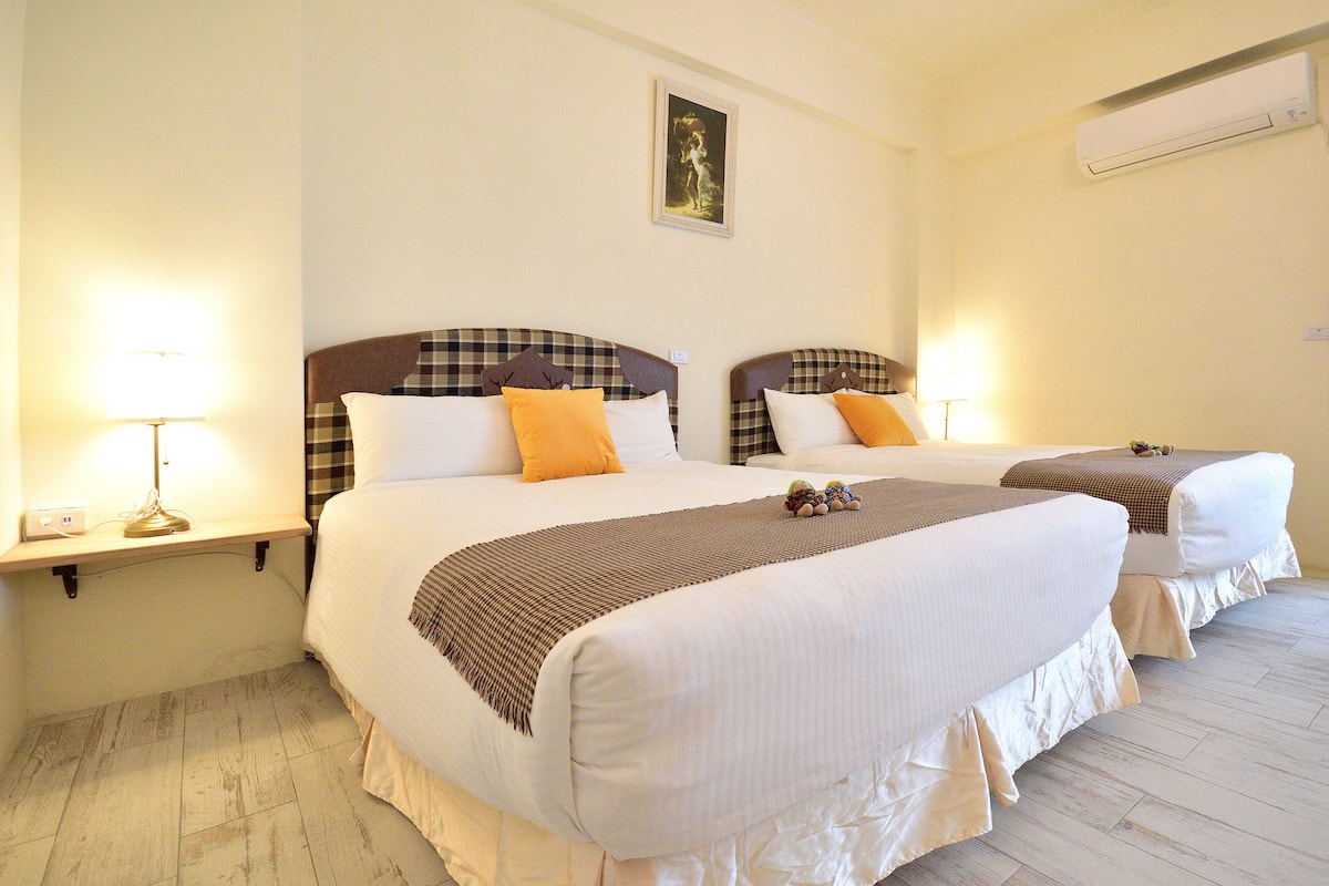 A hotel room with two beds, each with a headboard, bedside tables with lamps, and a wall-mounted air conditioner.