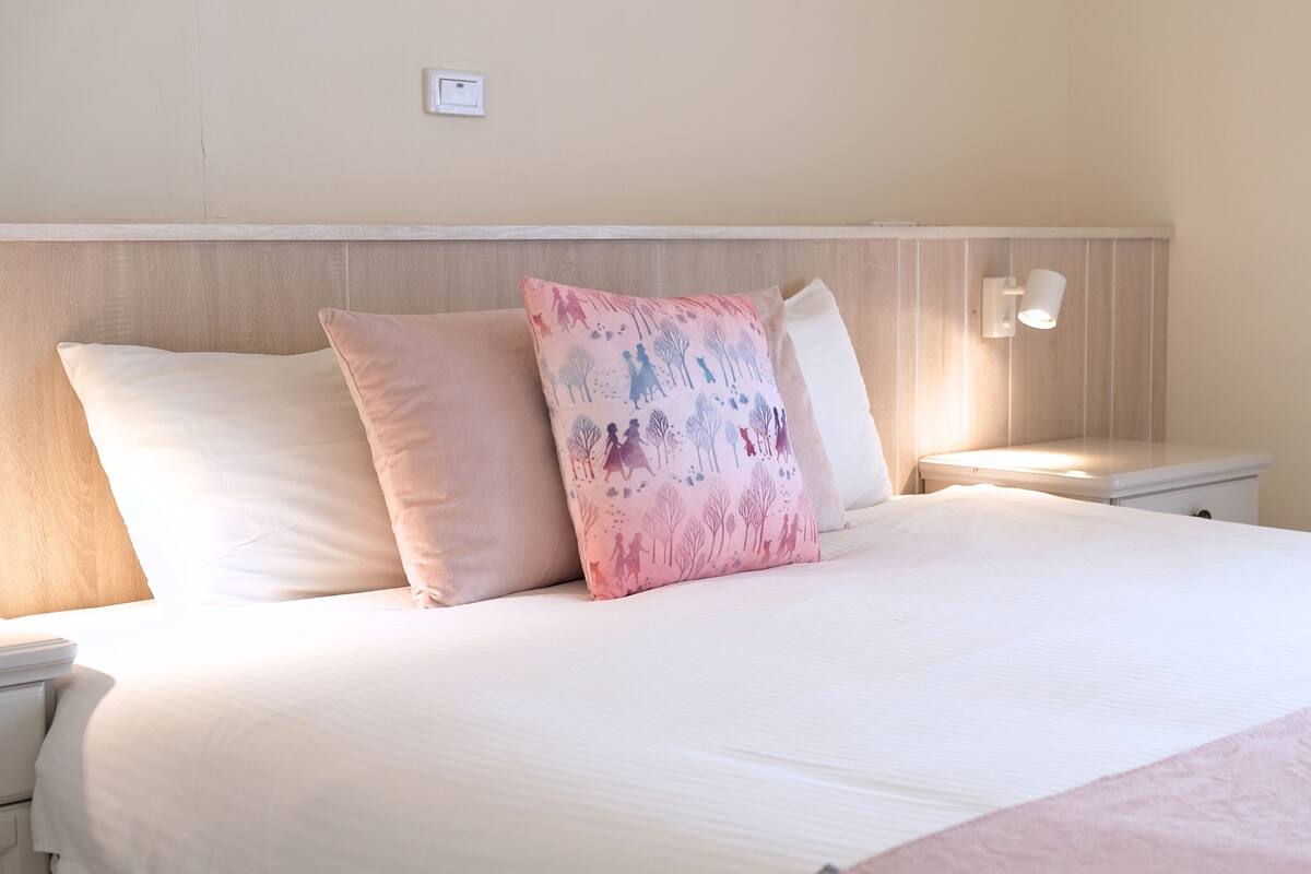 A neatly made bed with white bedding and three pillows, one with a tree pattern. There is a bedside lamp on the right and a small wall-mounted light above the bed.