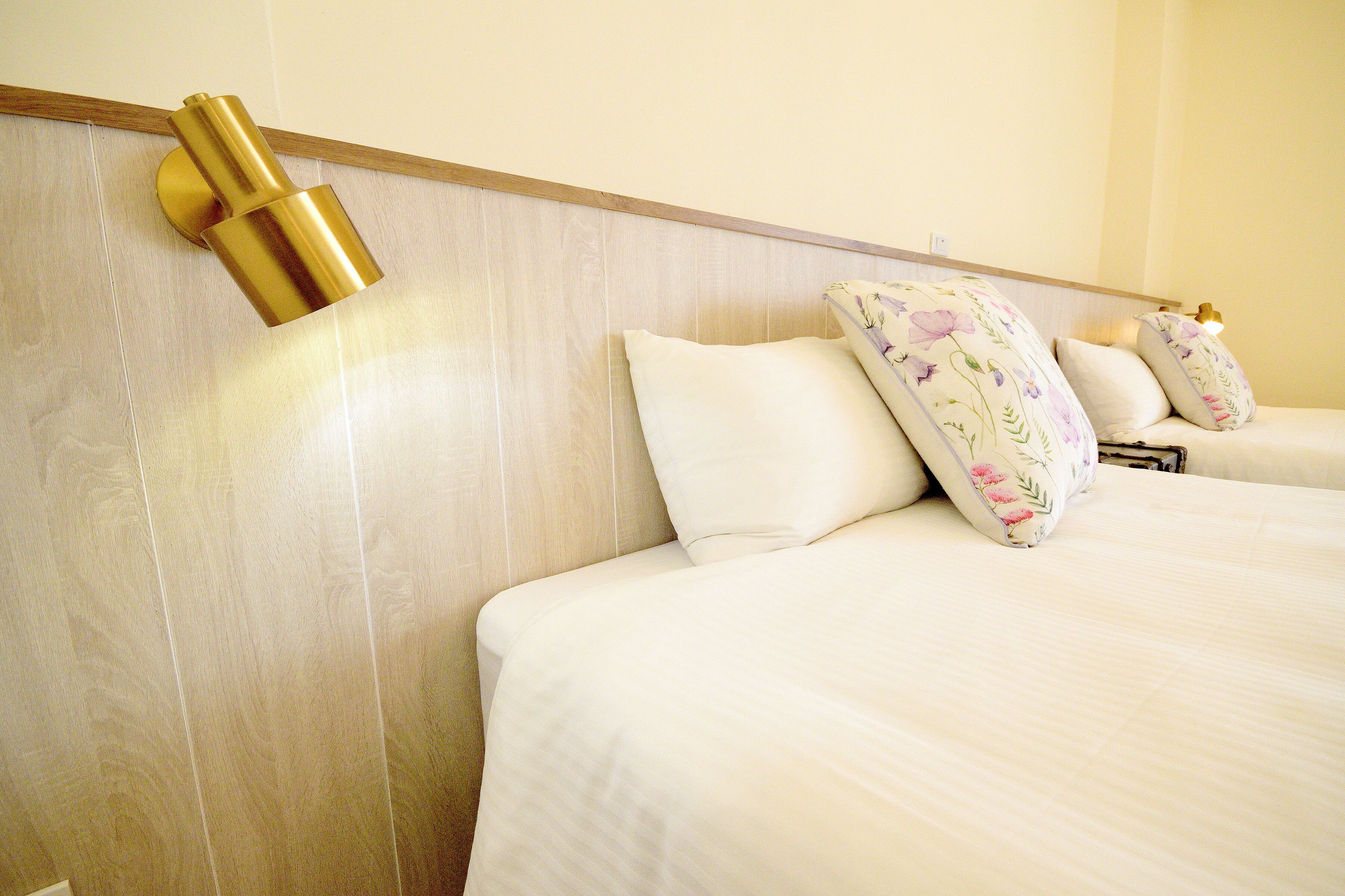 Habitación de hotel con cabecera de madera, dos camas con almohadas de estampado floral y una lámpara fijada a la pared.