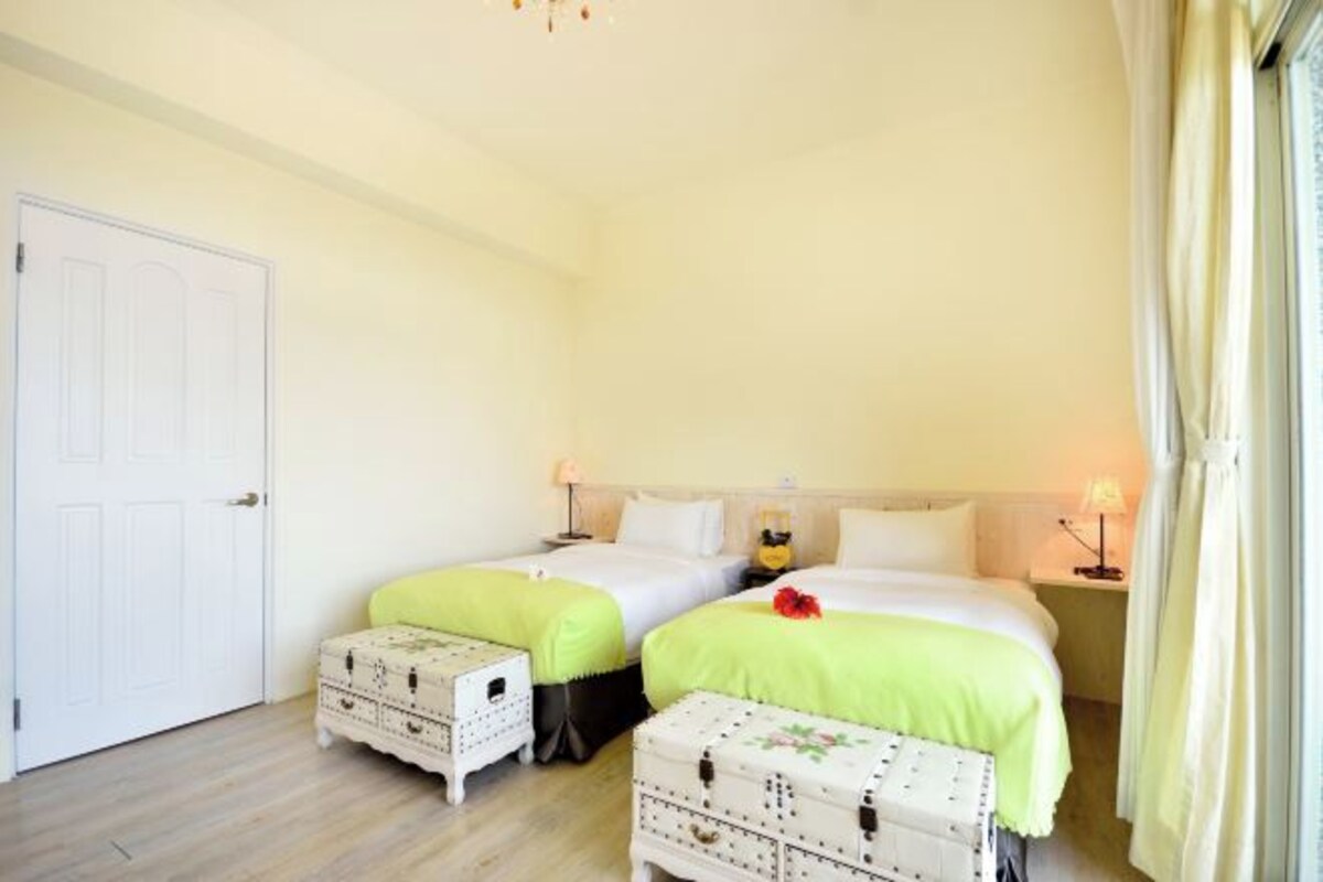 Une chambre avec deux lits, chacun recouvert d’une couverture verte et agrémenté d’oreillers blancs, et une porte blanche sur le côté gauche.