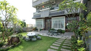 Edificio de dos pisos con un jardín que incluye un camino de piedra, una mesa redonda y plantas en macetas.
