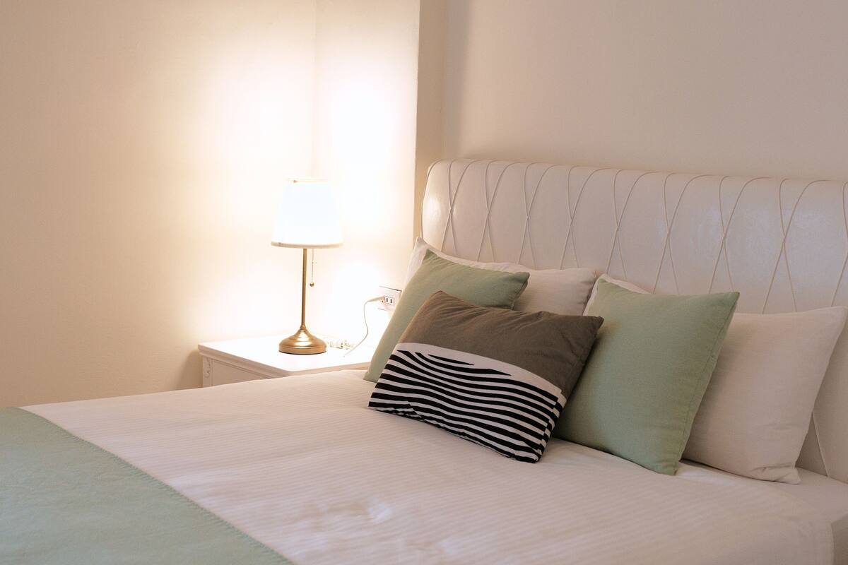 A neatly made bed with a striped pillow, a green pillow, and a white pillow. There is a bedside table with a lamp and a power outlet.