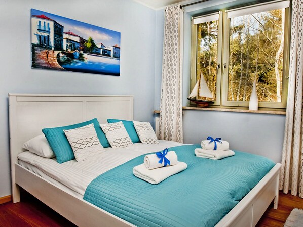 A neatly made bed with a teal quilt, white pillows, and a blue towel. A painting of a coastal scene hangs above the bed.