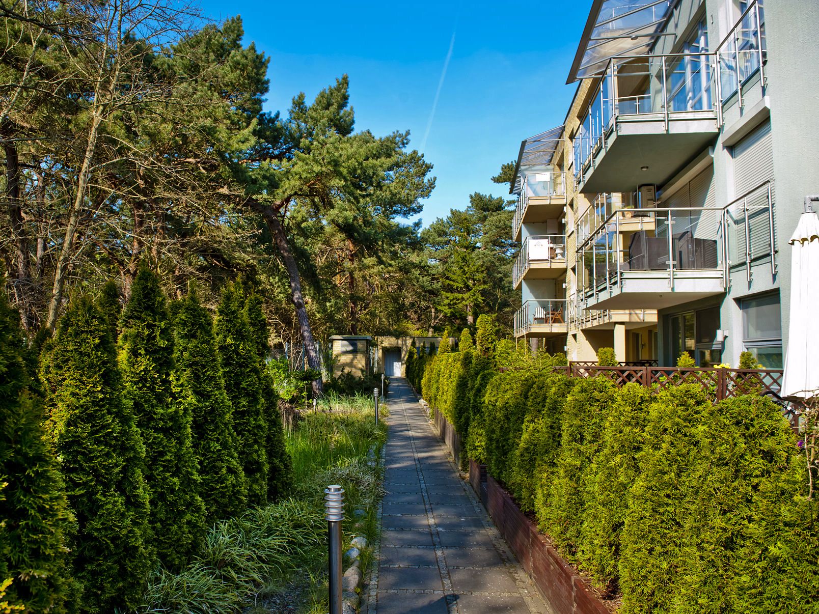 A pathway flanked by dense greenery and a multi-story building with balconies.