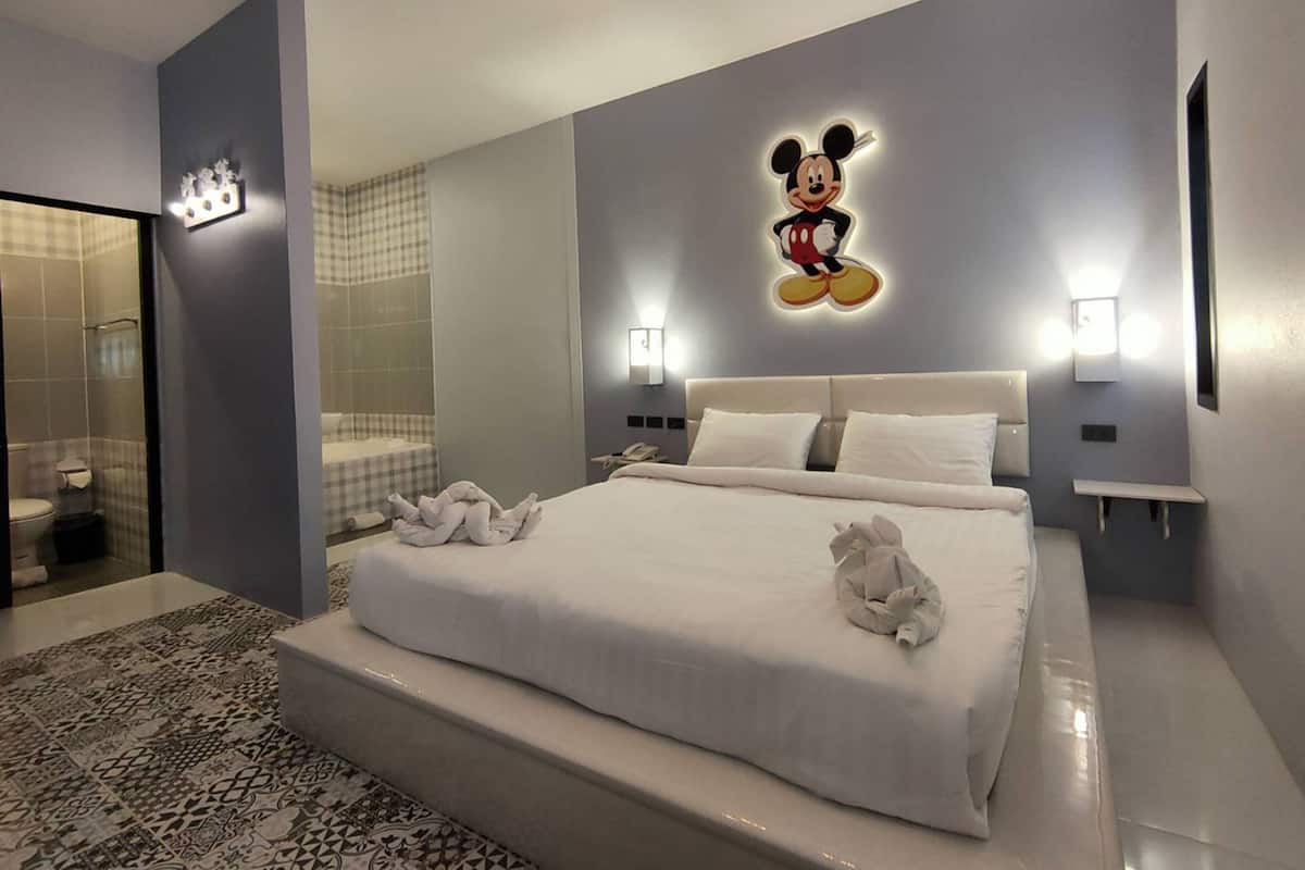 A modern hotel room with a large bed, a Mickey Mouse wall decoration, and a bathroom with a glass shower enclosure.