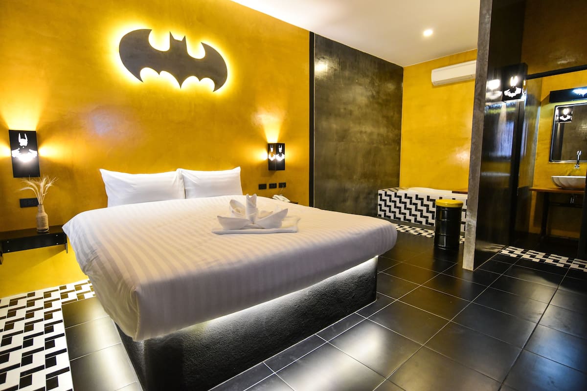 A hotel room with a bed, a wall-mounted Batman logo, a black and white tiled floor, and a bathroom with a sink and mirror.