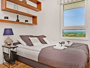 A neatly made bed with a brown blanket and pillows, a bedside table with a lamp, and a window with a view of the sea.