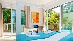 Ein modernes Hotelzimmer mit einem großen Bett, einem Schreibtisch mit einem Laptop und Blick auf Grün hinter großen Fenstern.