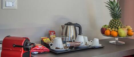 A breakfast setup with a red coffee machine, a silver kettle, white cups, a tray with pastries, a fruit bowl, and a bag of coffee.