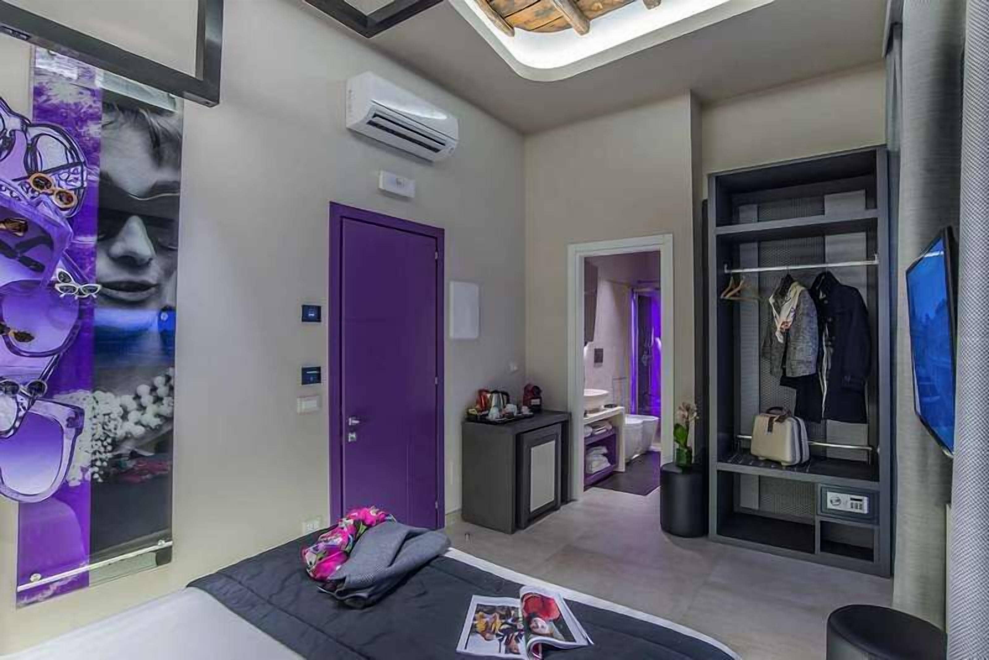 Ein modernes Hotelzimmer mit violetter Tür, einem Bett mit einer Zeitschrift, einem kleinen Tisch mit einer Lampe, einem Kleiderschrank mit Kleidung und einem Fernseher an der Wand.