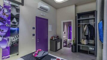 Ein modernes Hotelzimmer mit violetter Tür, einem Bett mit einer Zeitschrift, einem kleinen Tisch mit einer Lampe, einem Kleiderschrank mit Kleidung und einem Fernseher an der Wand.