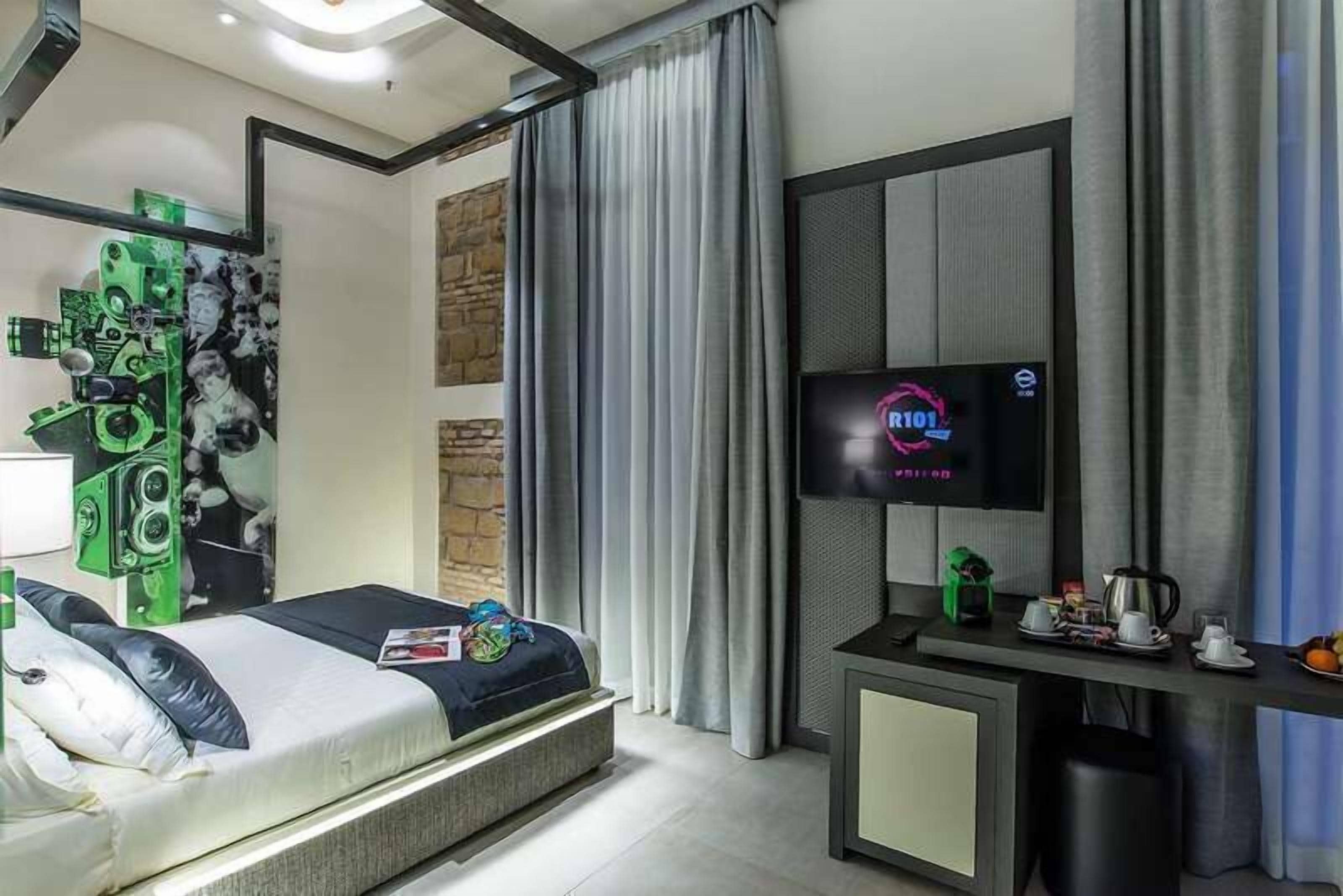 Una habitación de hotel moderna con una cama, un televisor, un escritorio con cafetera y una pared con un diseño abstracto en tonos verde y negro.