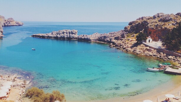 A coastal area with clear turquoise waters, rocky terrain, and a small white building near the shore. - Residenza Maria Lindos Villa (Rhodes)
